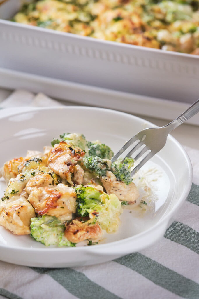 A fork holding a bite of broccoli and chicken above a white plate filled with a chicken and broccoli dish.
