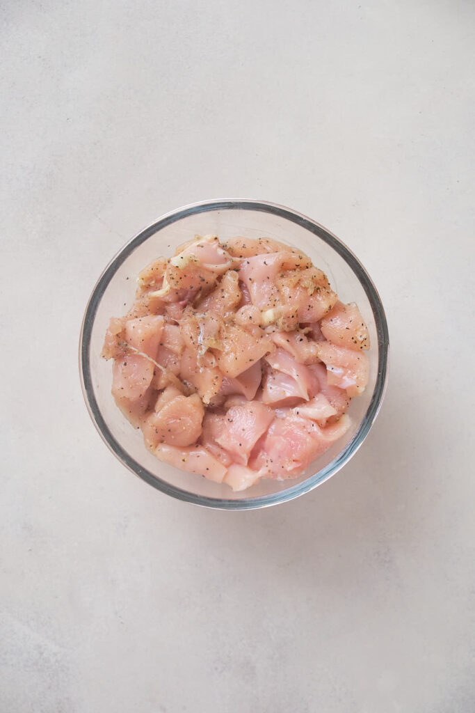A glass bowl filled with raw, seasoned chicken pieces on a light-colored surface.