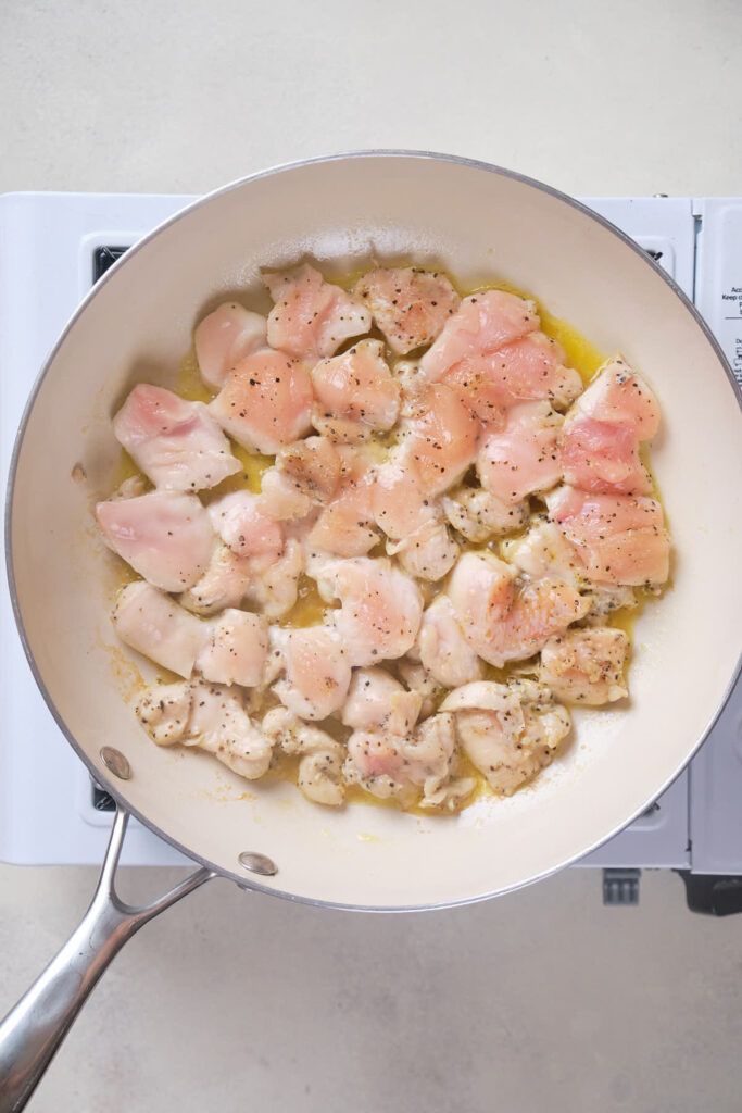 Diced chicken pieces are being cooked in a white frying pan on a stovetop.