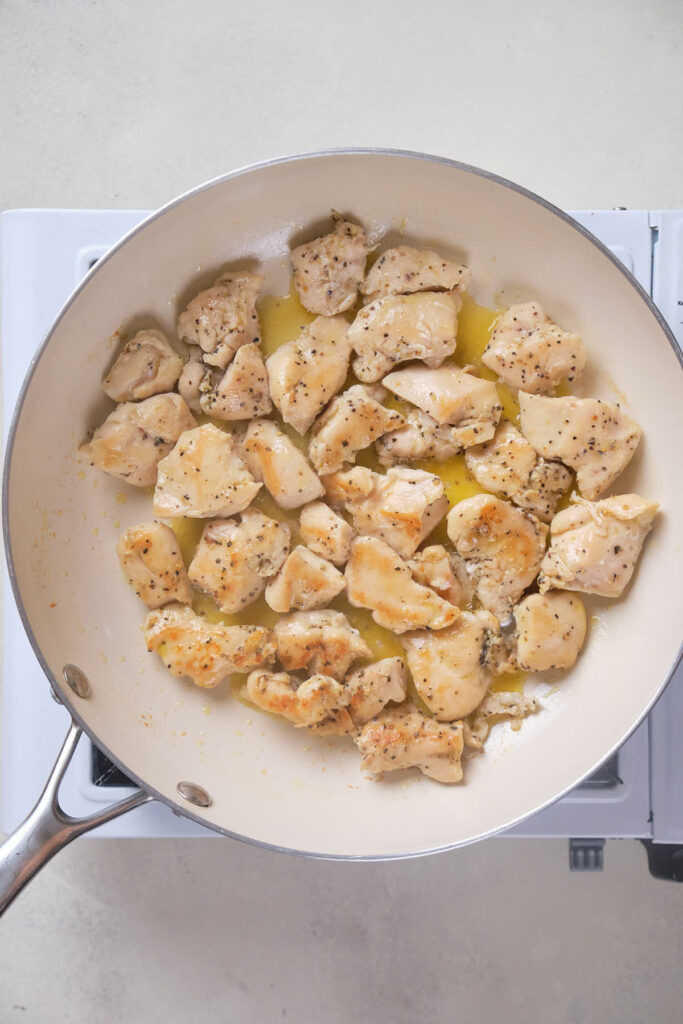 Cooked, diced chicken pieces in a white frying pan on a stovetop, seasoned with pepper and lightly browned.