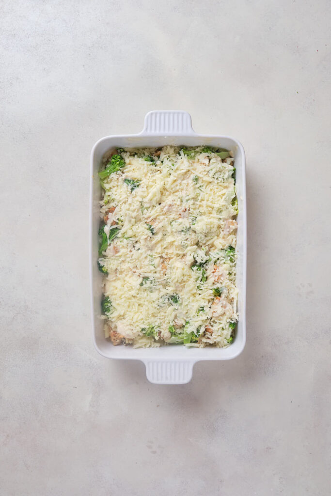 A white rectangular baking dish filled with a mixture of shredded cheese, chopped broccoli, and small pieces of chicken.