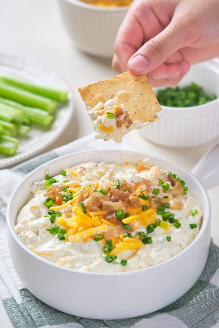 A hand holding a cracker dipped in a white bowl of creamy dip topped with cheese and bacon.