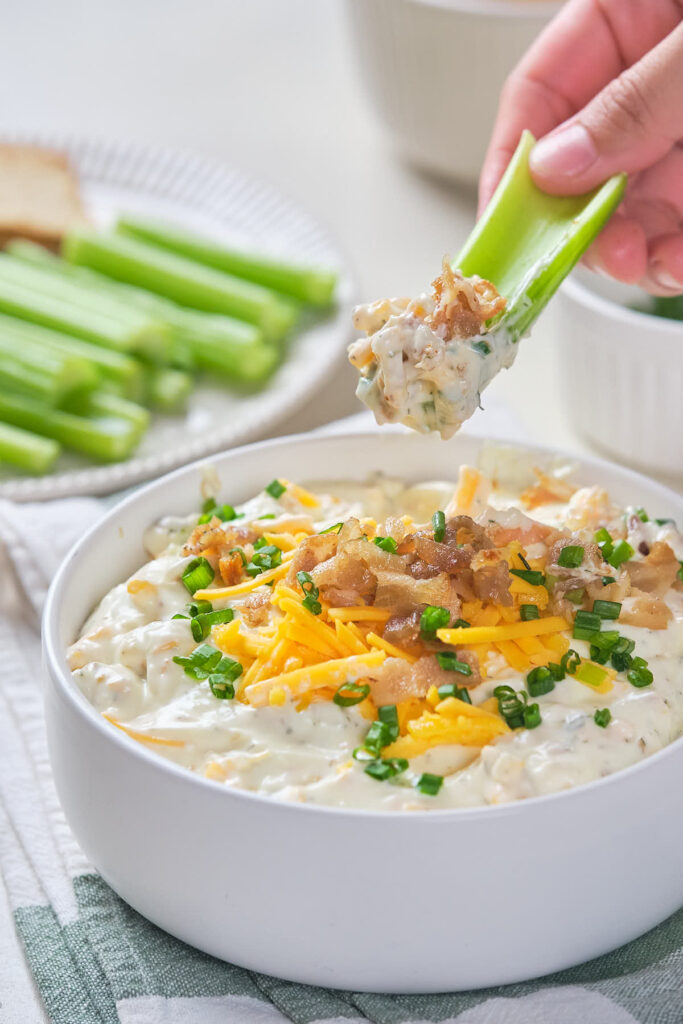 A hand dipping a celery stick into a bowl of creamy dip topped with cheese.