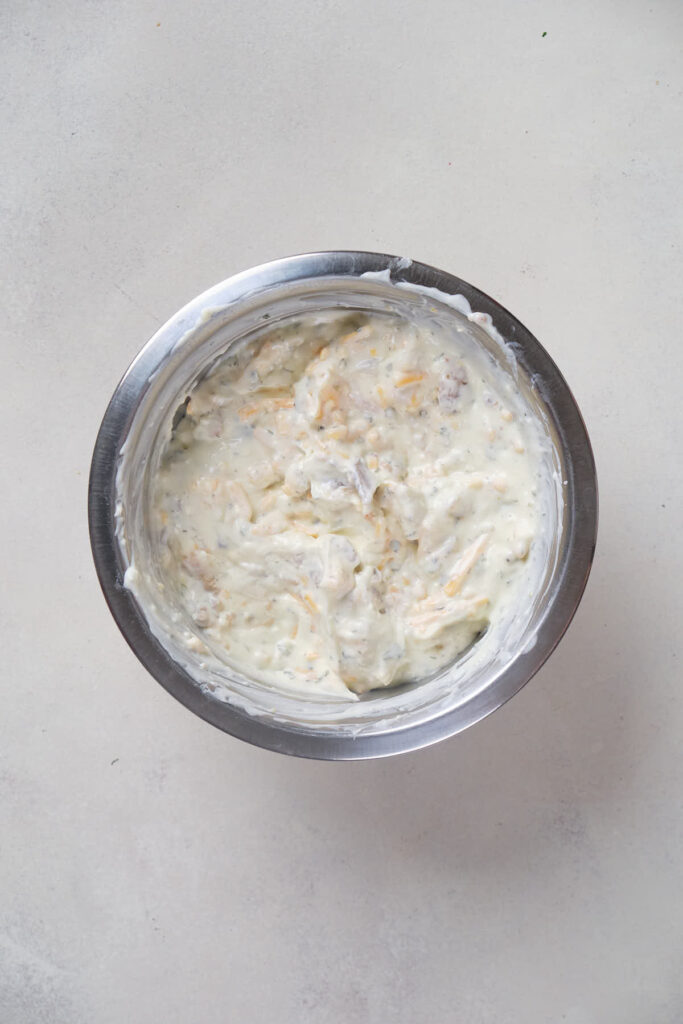 A bowl of creamy dip with visible herbs and shredded ingredients, placed on a light-colored surface.