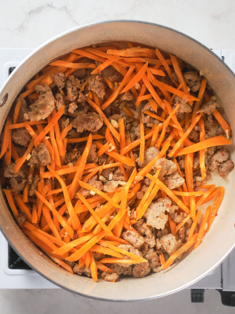 A pot on a stovetop contains ground meat and thinly sliced carrots being cooked together.