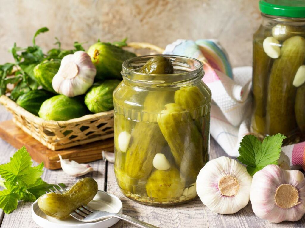 A jar of pickles with garlic sits on a table near a basket of fresh cucumbers and garlic cloves.