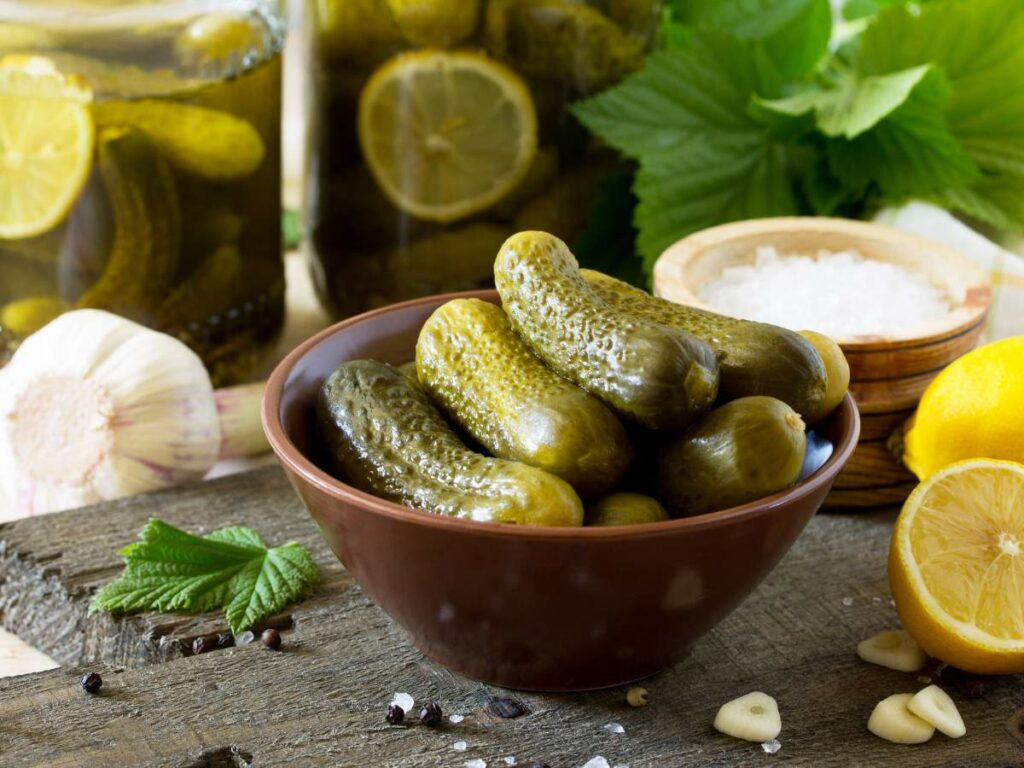 A bowl of pickles on a wooden table, surrounded by garlic, lemon halves, salt, and jars of pickles in the background. There are also green leaves and black peppercorns scattered around.