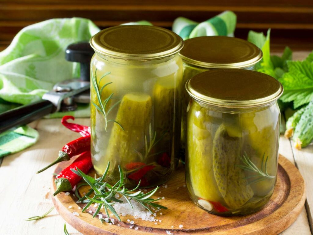 Three jars of pickles with red chili peppers and rosemary, placed on a wooden board with a can opener and green cloth in the background.