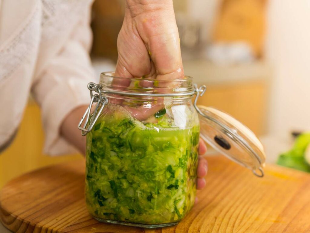 A hand is pressing down cabbage inside a glass jar on a wooden surface.