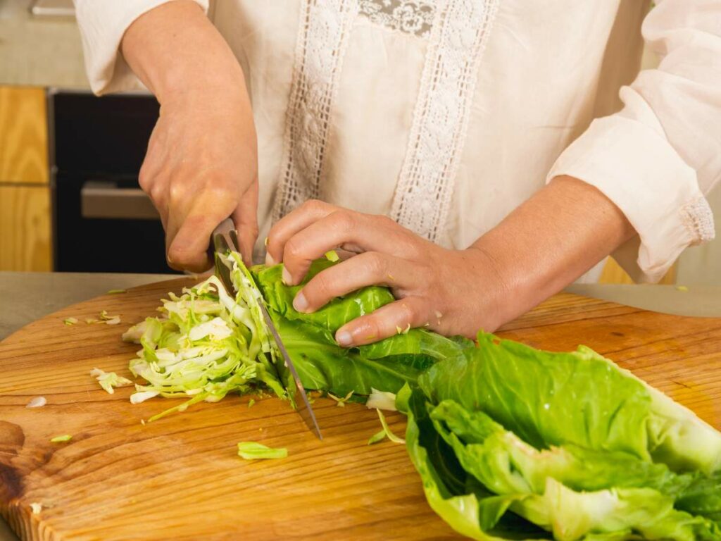 Person slicing cabbage on a wooden cutting board with a knife in a kitchen.