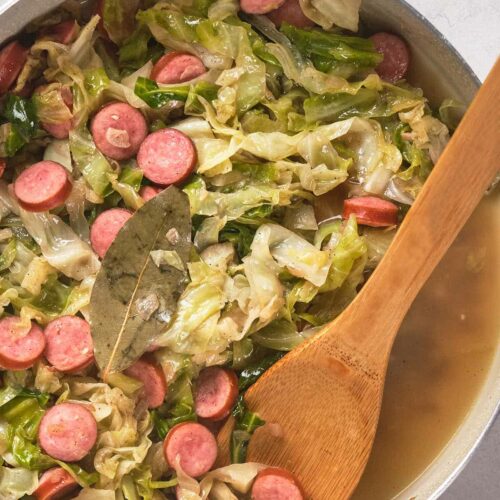 A pot filled with cooked cabbage, sliced sausages, and broth, garnished with a bay leaf.