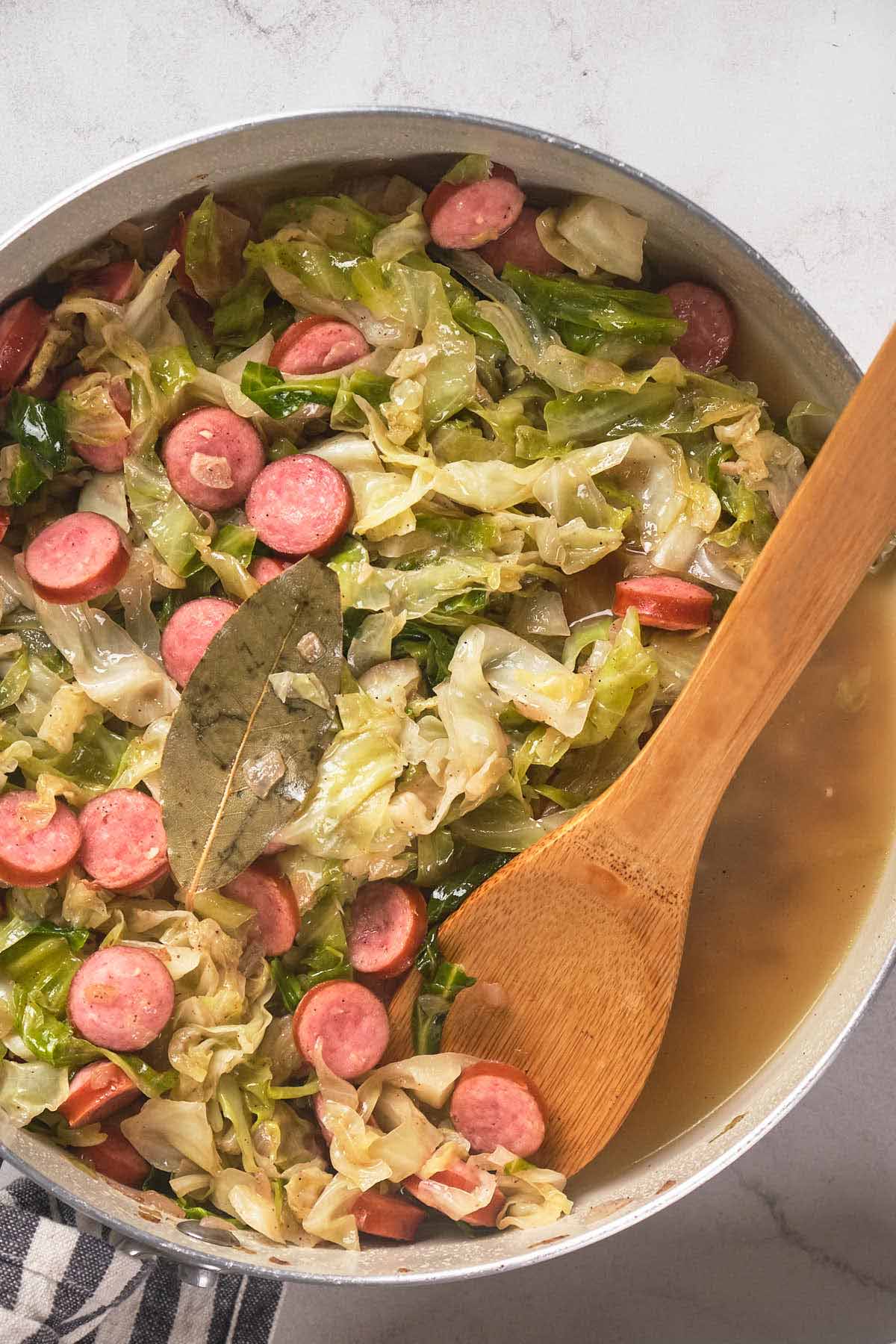 A pot filled with cooked cabbage, sliced sausages, and broth, garnished with a bay leaf.
