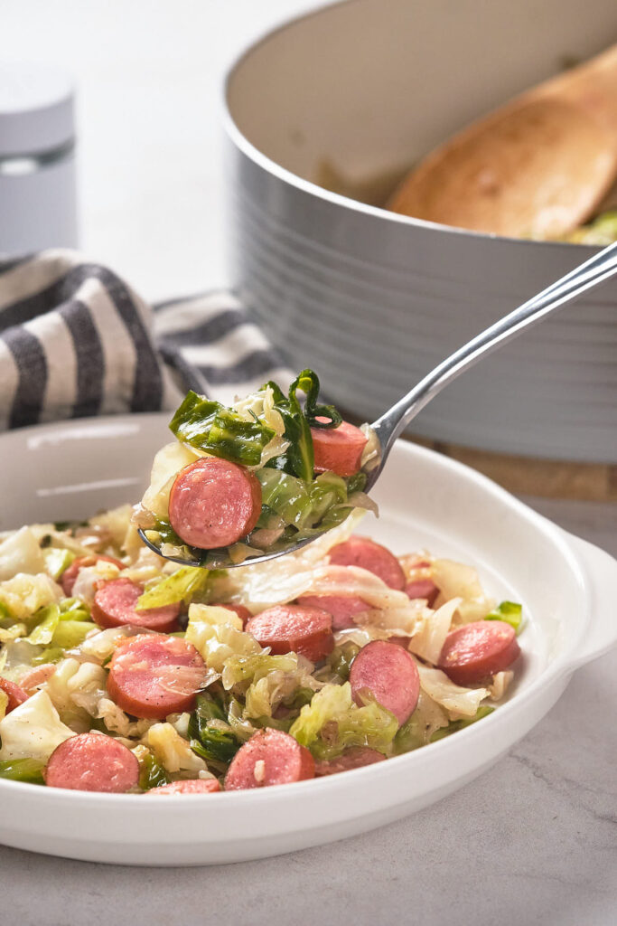 A spoonful of sausage and cabbage is held above a plate filled with the same dish.