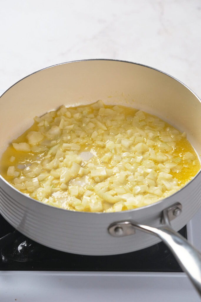 A large saucepan on a stove with diced onions being sautéed in melted butter.