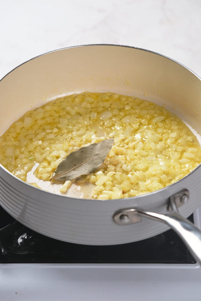 A saucepan with chopped onions and a bay leaf cooking in oil on a stove.