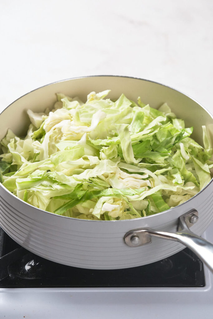 A close-up of a pan filled with chopped green cabbage, cooking on a stove.