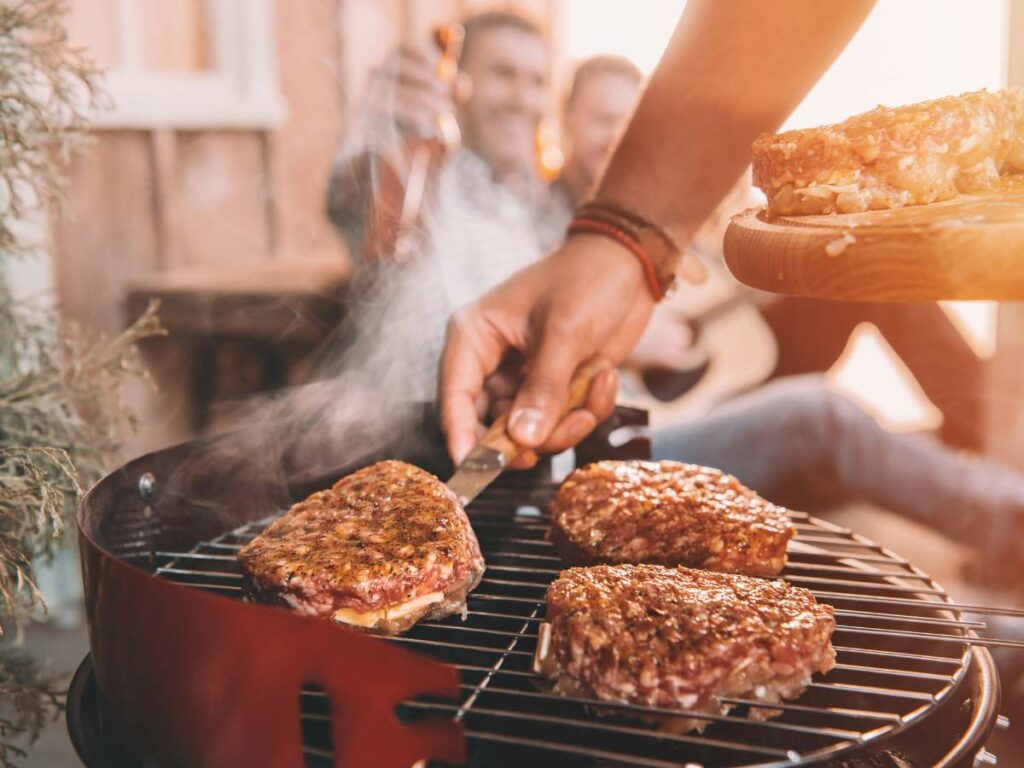 A person is grilling steaks on a barbecue grill. Steam is rising from the steaks, and three people can be seen in the background, one holding a drink and the other two sitting.