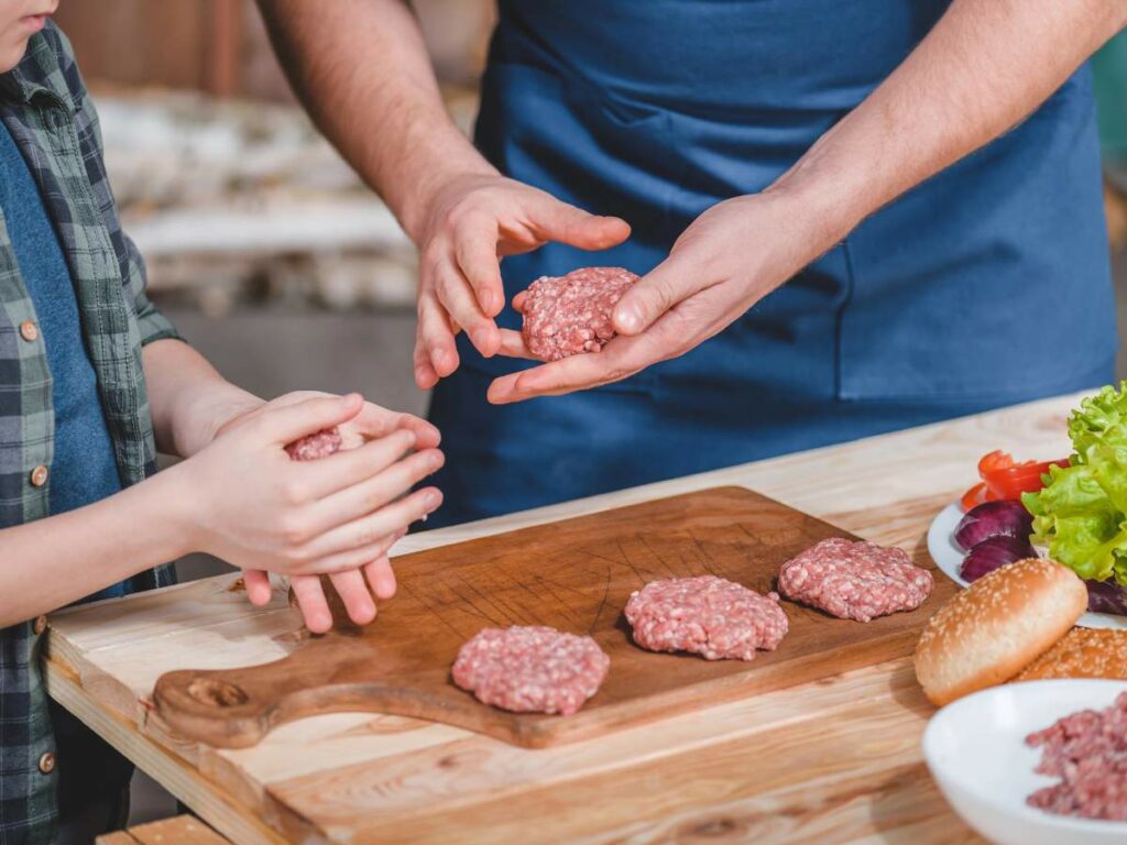 Two people shape raw burger patties by hand on a wooden cutting board, preparing ingredients for burgers, which include lettuce, red onion, and a burger bun.