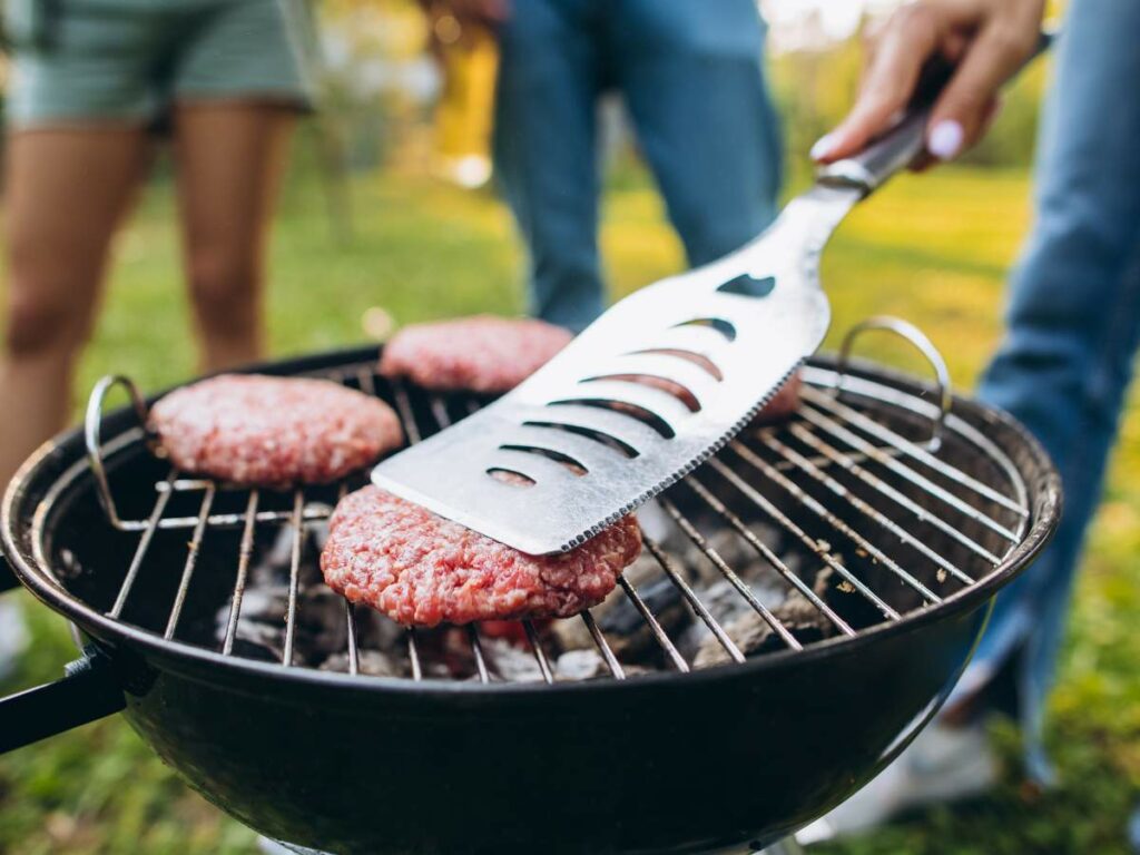 Three people are grilling hamburger patties on a charcoal grill. One person holds a spatula, flipping a patty. The scene takes place outdoors in a grassy area.