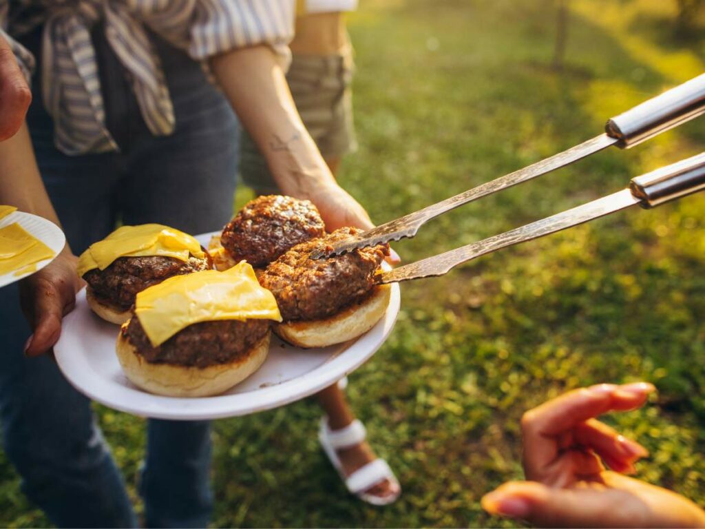 Close-up of a person holding a plate with three cheeseburgers being placed on buns using skewers at an outdoor gathering.