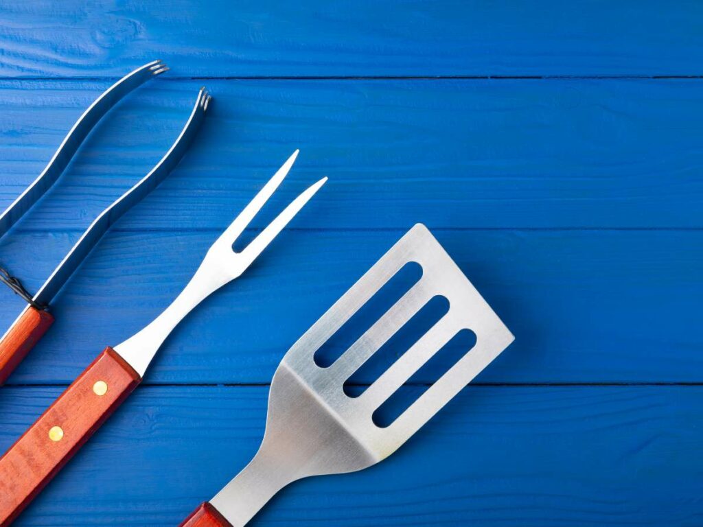 Three barbecue tools with wooden handles—a fork, a spatula, and tongs—laid out on a blue wooden surface.