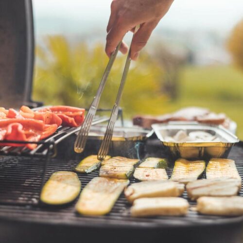 A person uses tongs to grill vegetables and other foods on an outdoor barbecue grill on a sunny day.