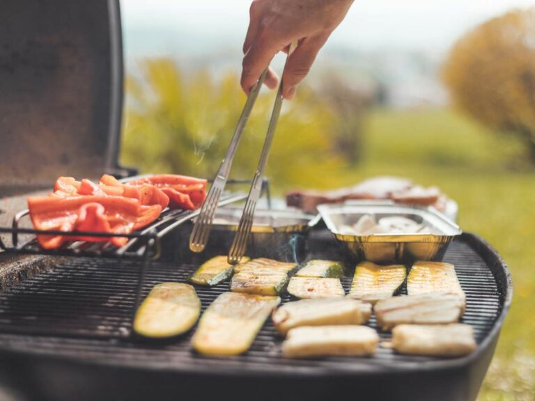 A person uses tongs to grill vegetables and other foods on an outdoor barbecue grill on a sunny day.