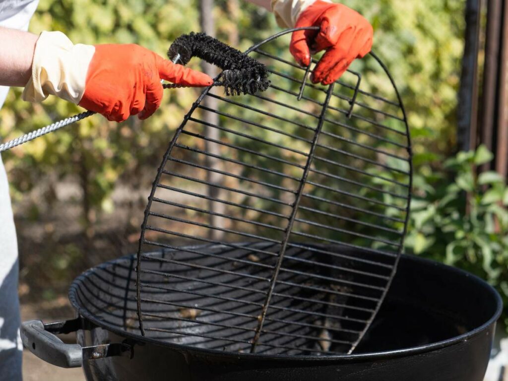 A person wearing red gloves is scrubbing a black grill grate with a wire brush over a round barbecue grill outdoors.