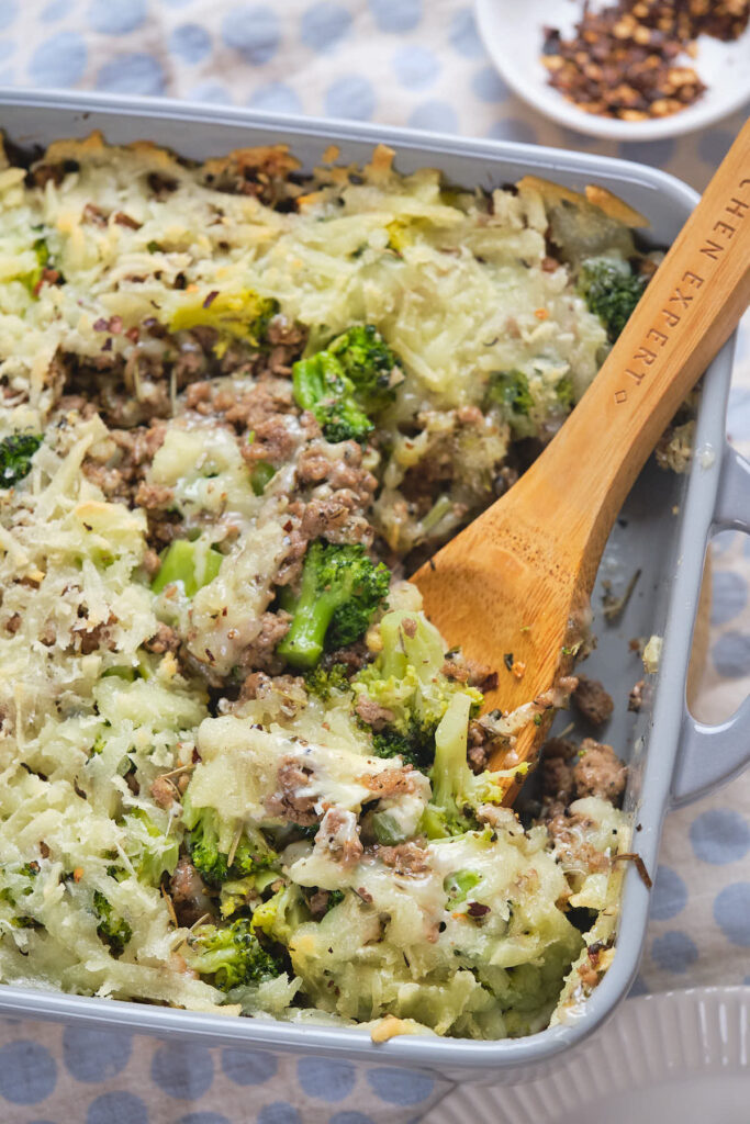 A casserole dish with a wooden spoon, featuring a mixture of cooked ground meat, broccoli, and melted cheese. A small bowl of red pepper flakes is in the background.