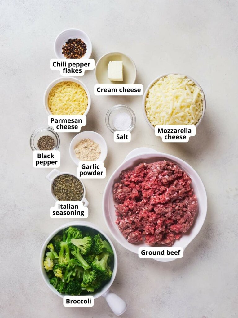 Ingredients for a recipe arranged on a countertop: chili pepper flakes, cream cheese, Parmesan cheese, mozzarella cheese, black pepper, salt, garlic powder, Italian seasoning, ground beef, and broccoli.