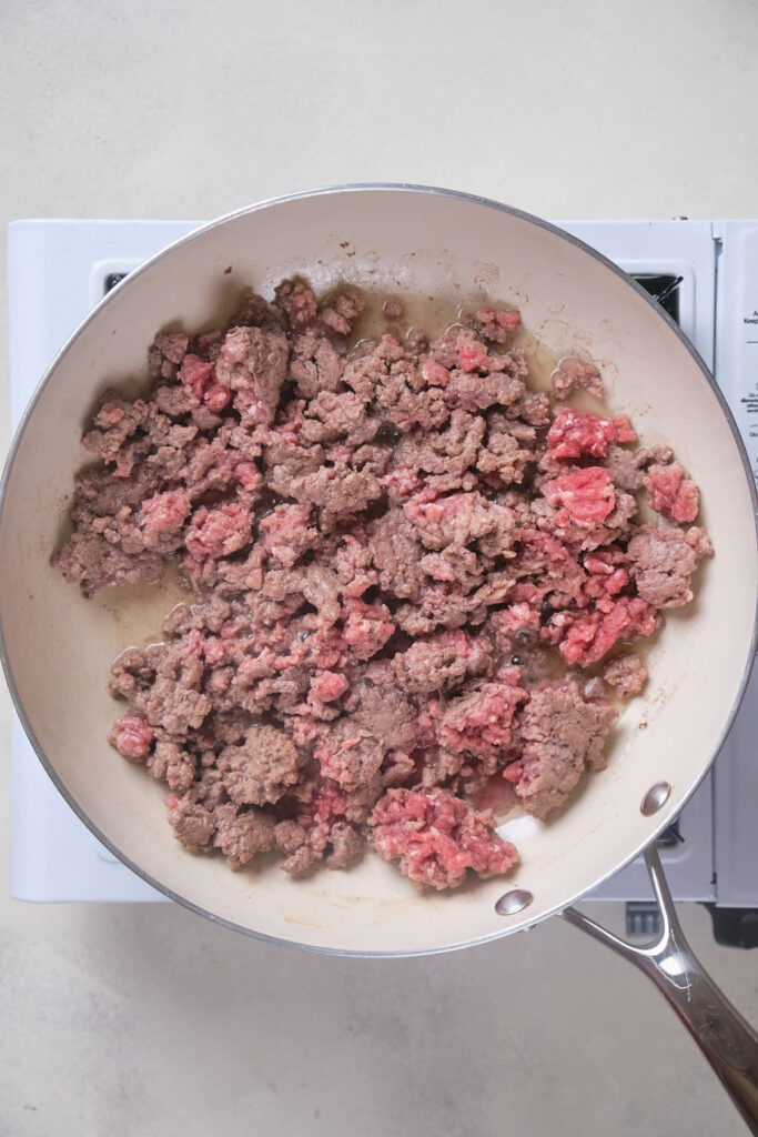 A frying pan on a stove contains partially cooked ground beef.