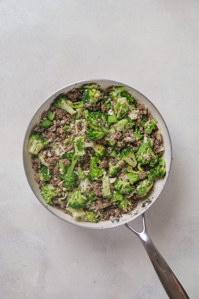 A frying pan filled with cooked ground meat and broccoli on a plain, light-colored surface.