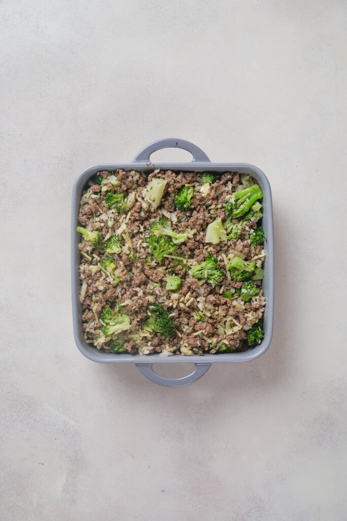 A square baking dish containing a mixture of cooked ground beef and broccoli on a light-colored surface.