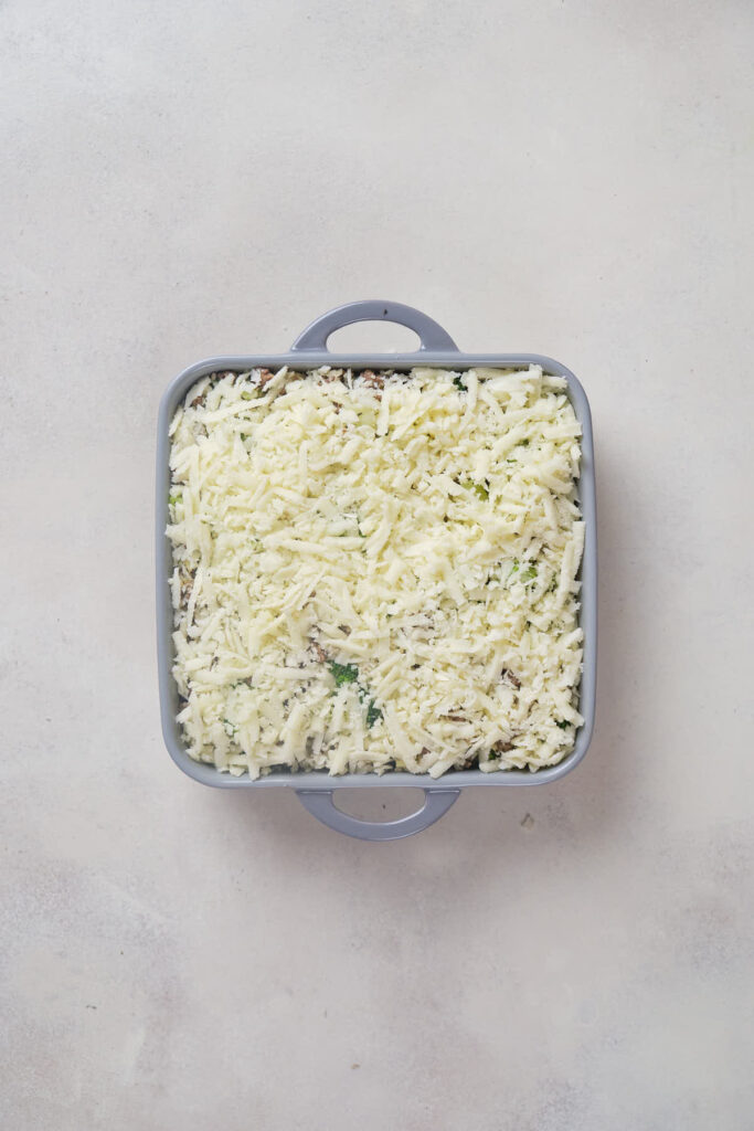 A square baking dish filled with a mixture topped with grated cheese on a light-colored surface.