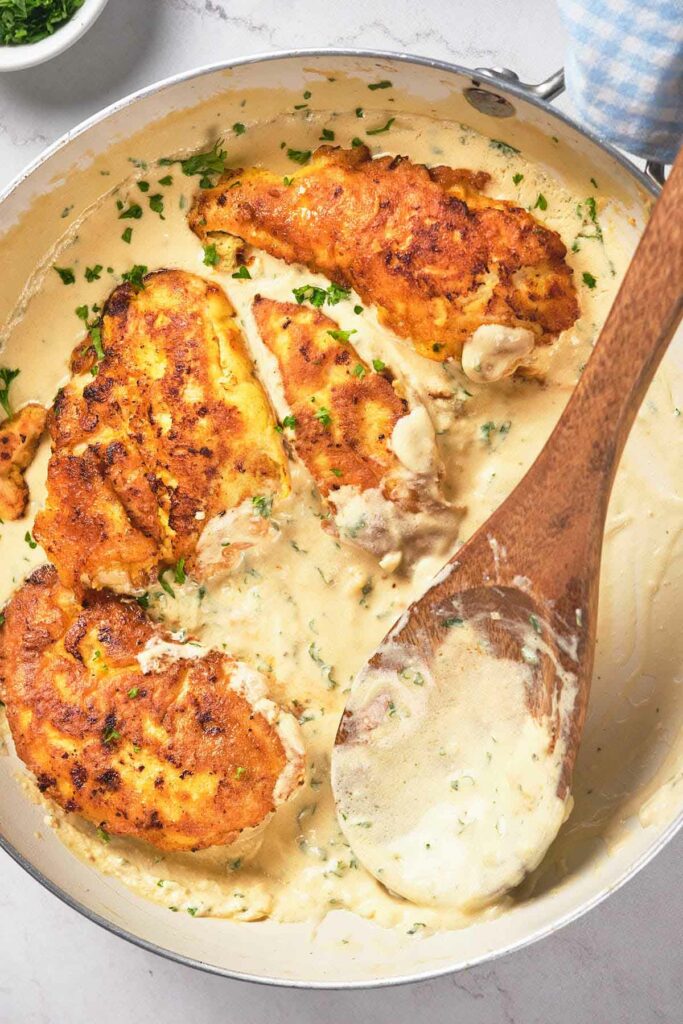 A skillet contains seasoned chicken breasts in a creamy sauce, garnished with chopped herbs. A wooden spoon rests inside the skillet, partially submerged in the sauce.
