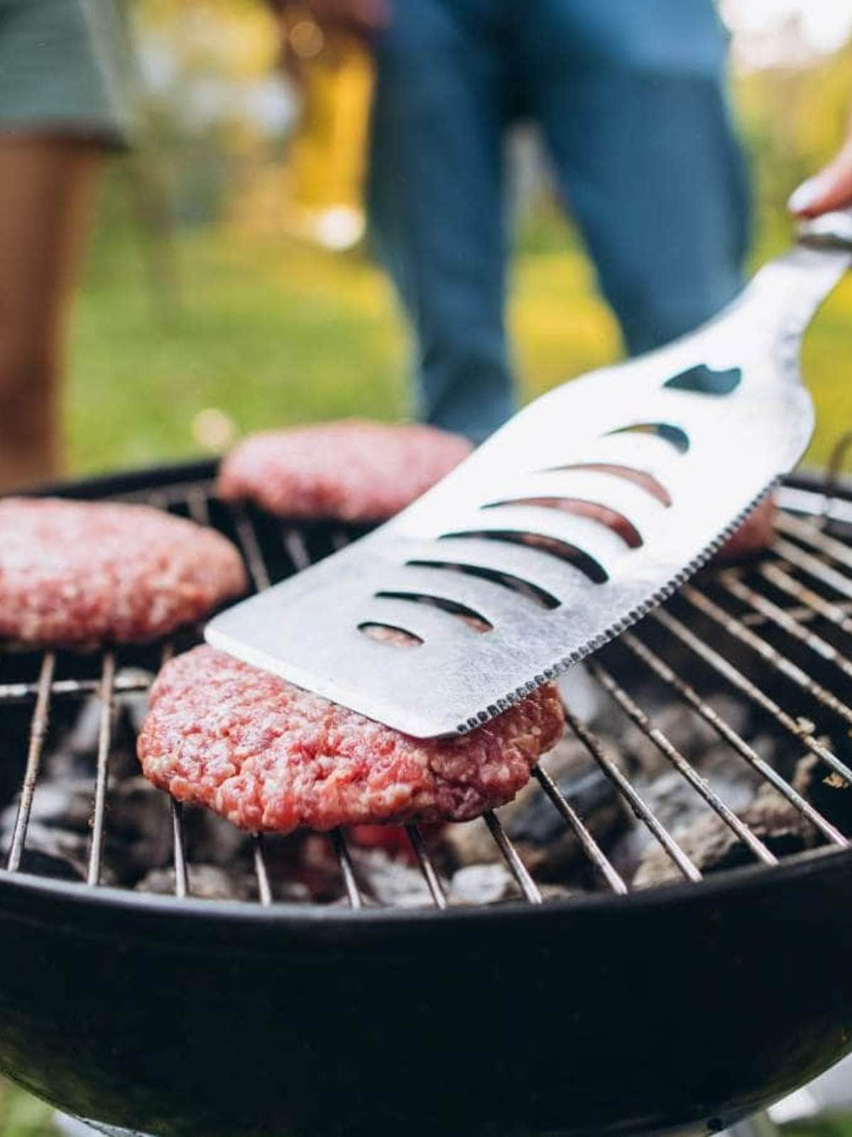 A metal spatula presses down on raw burger patties cooking on a round charcoal grill.