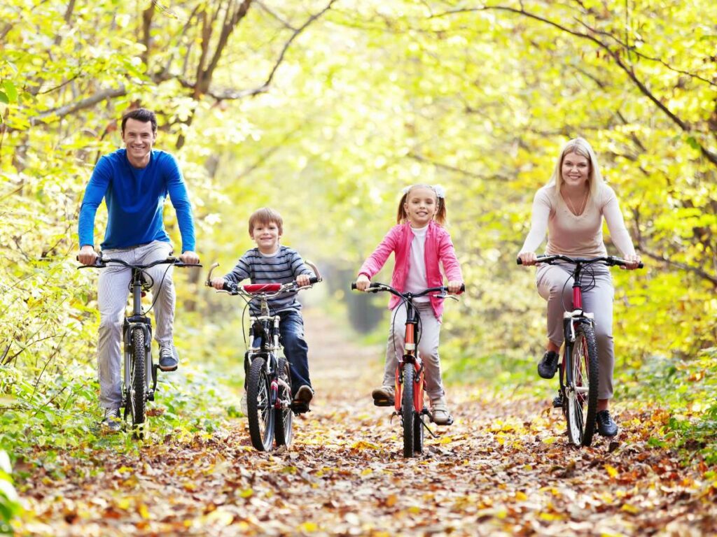 A family of four rides bicycles on a leafy path surrounded by trees in autumn.