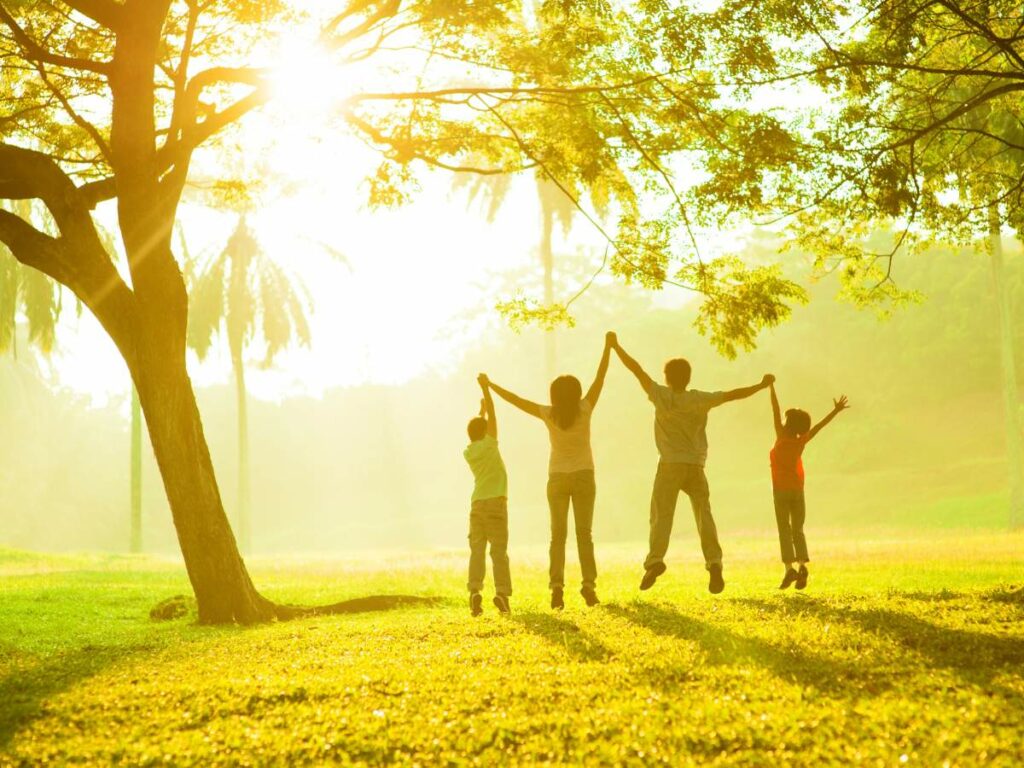 A family of four holding hands and jumping beneath a tree in a sunlit park.