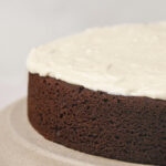 A close-up of a round chocolate cake with white frosting on top, placed on a beige cake stand.