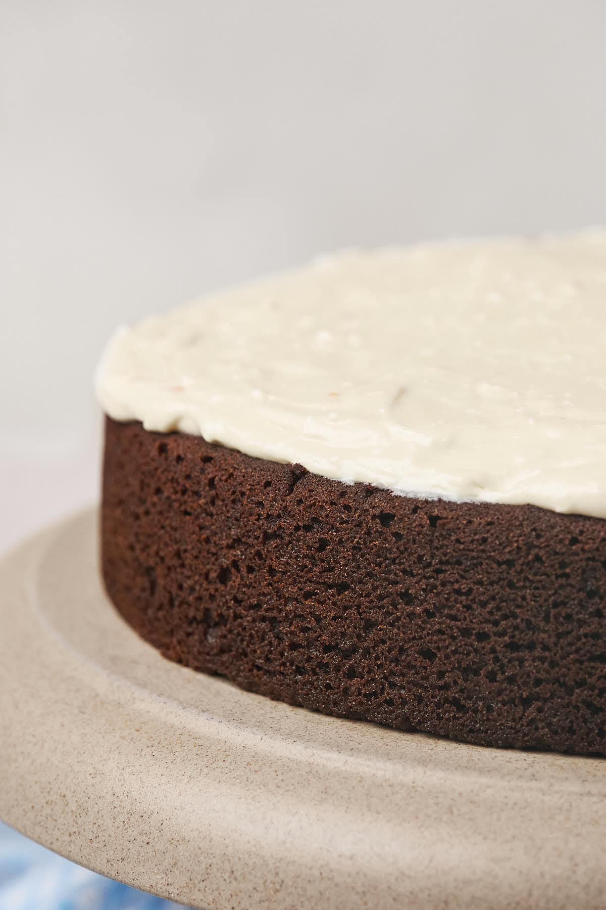 A close-up of a round chocolate cake with white frosting on top, placed on a beige cake stand.