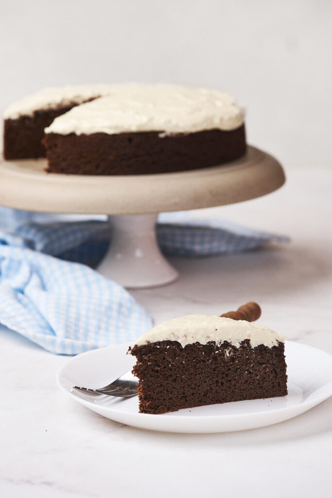 A slice of chocolate cake with white frosting is served on a plate with a fork in the foreground.