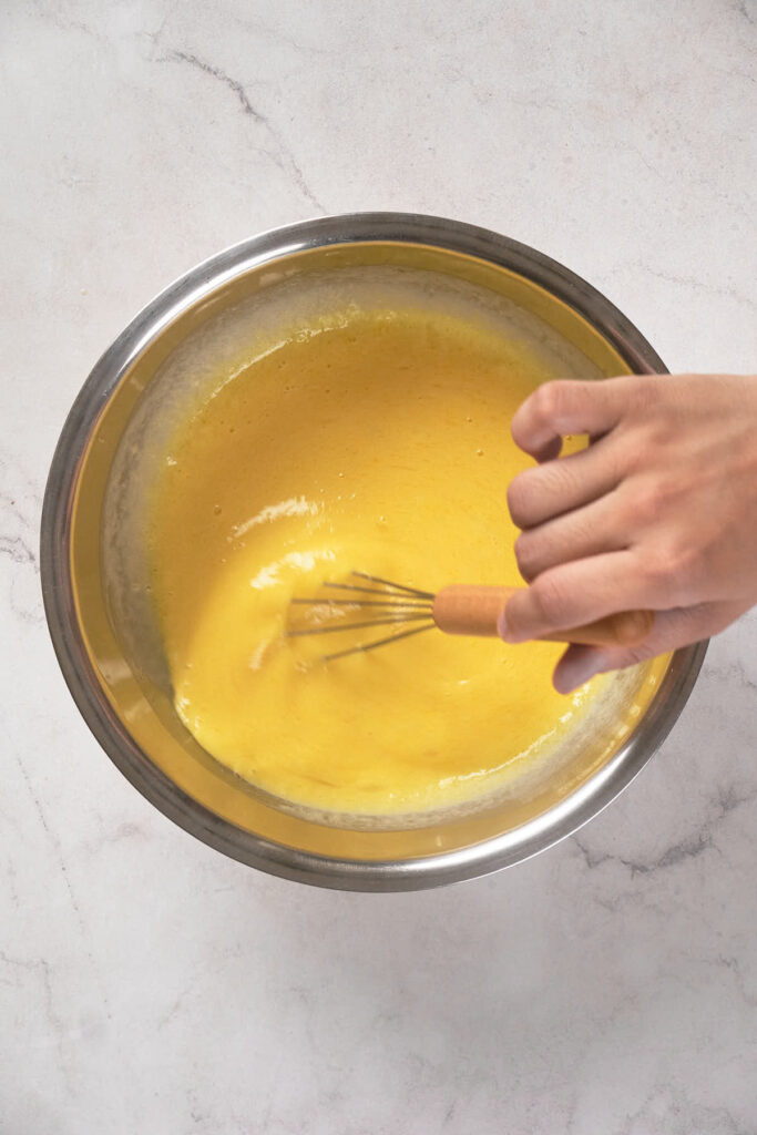 A hand whisking a yellow mixture in a stainless steel bowl on a white marble surface.