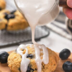 Close-up of a hand pouring white icing over a freshly baked blueberry scone.