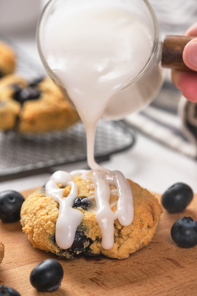 Close-up of a hand pouring white icing over a freshly baked blueberry scone.