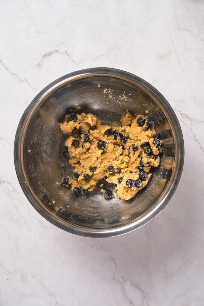 A stainless steel mixing bowl containing partially mixed scone dough with visible blueberries.