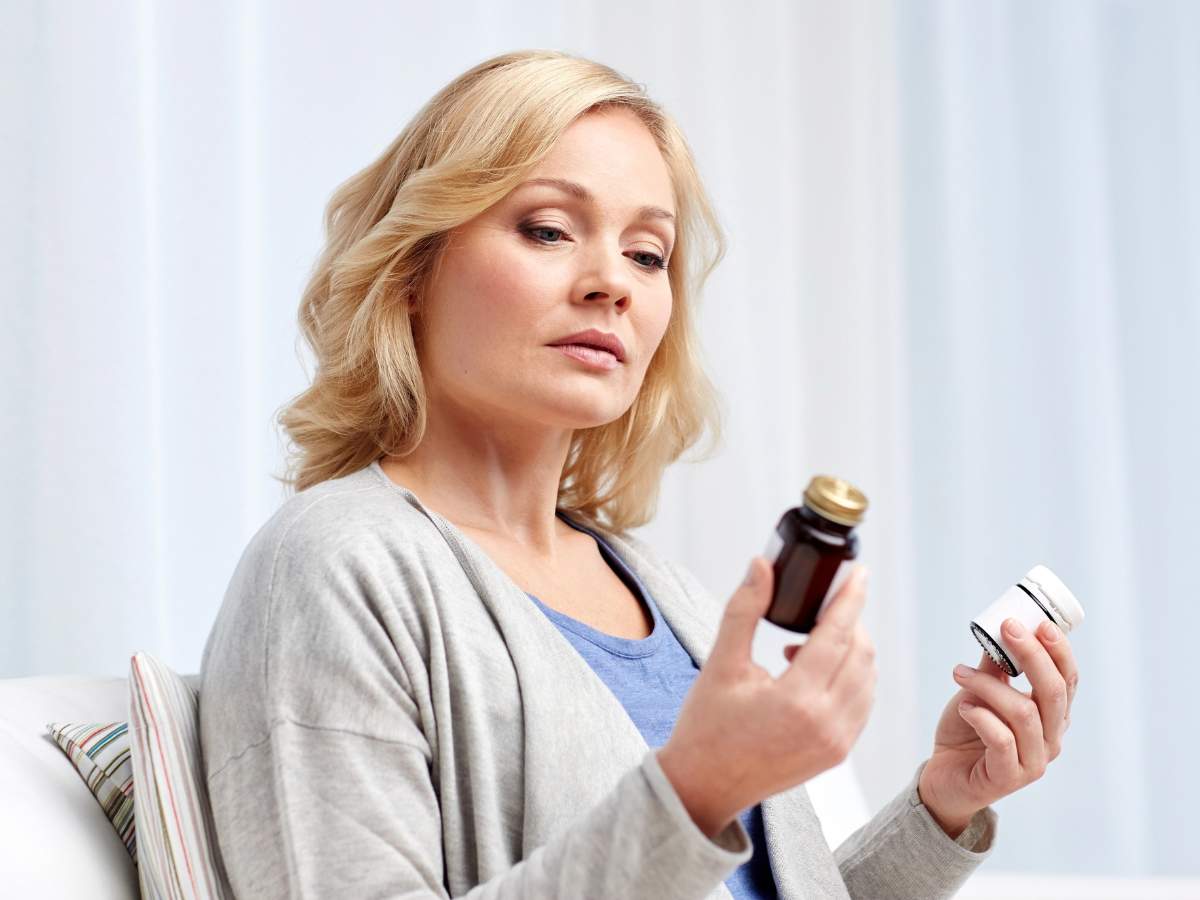 A woman with blonde hair examines a bottle of medication in one hand and holds another container in the other hand while seated indoors.