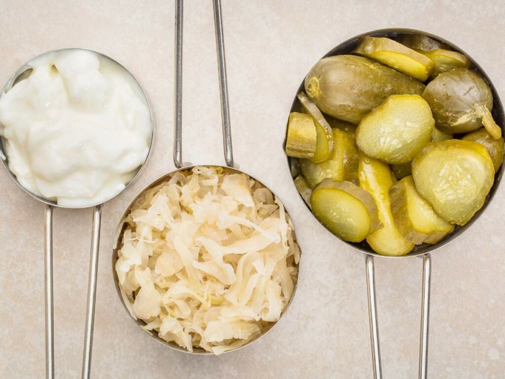 Three measuring cups on a beige surface, containing Greek yogurt, sauerkraut, and pickles from left to right.