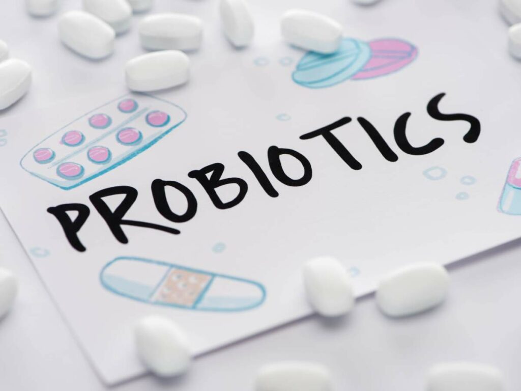 The word "PROBIOTICS" is written on a piece of paper surrounded by illustrations of pills, capsules, and tablets, with white pills scattered around.