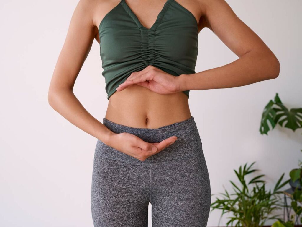 A person in a green top and gray leggings stands with their hands gesturing around their midsection. Plants are visible in the background.