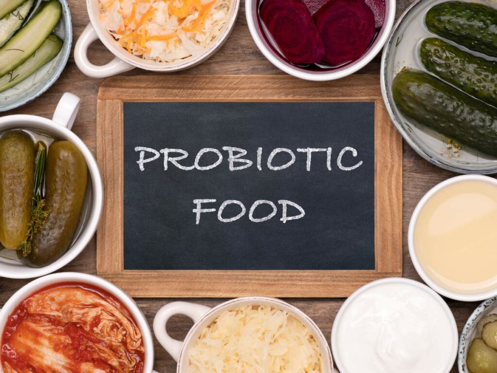 A chalkboard labeled "Probiotic Food" surrounded by various fermented foods including pickles, kimchi, yogurt, and sauerkraut in bowls.